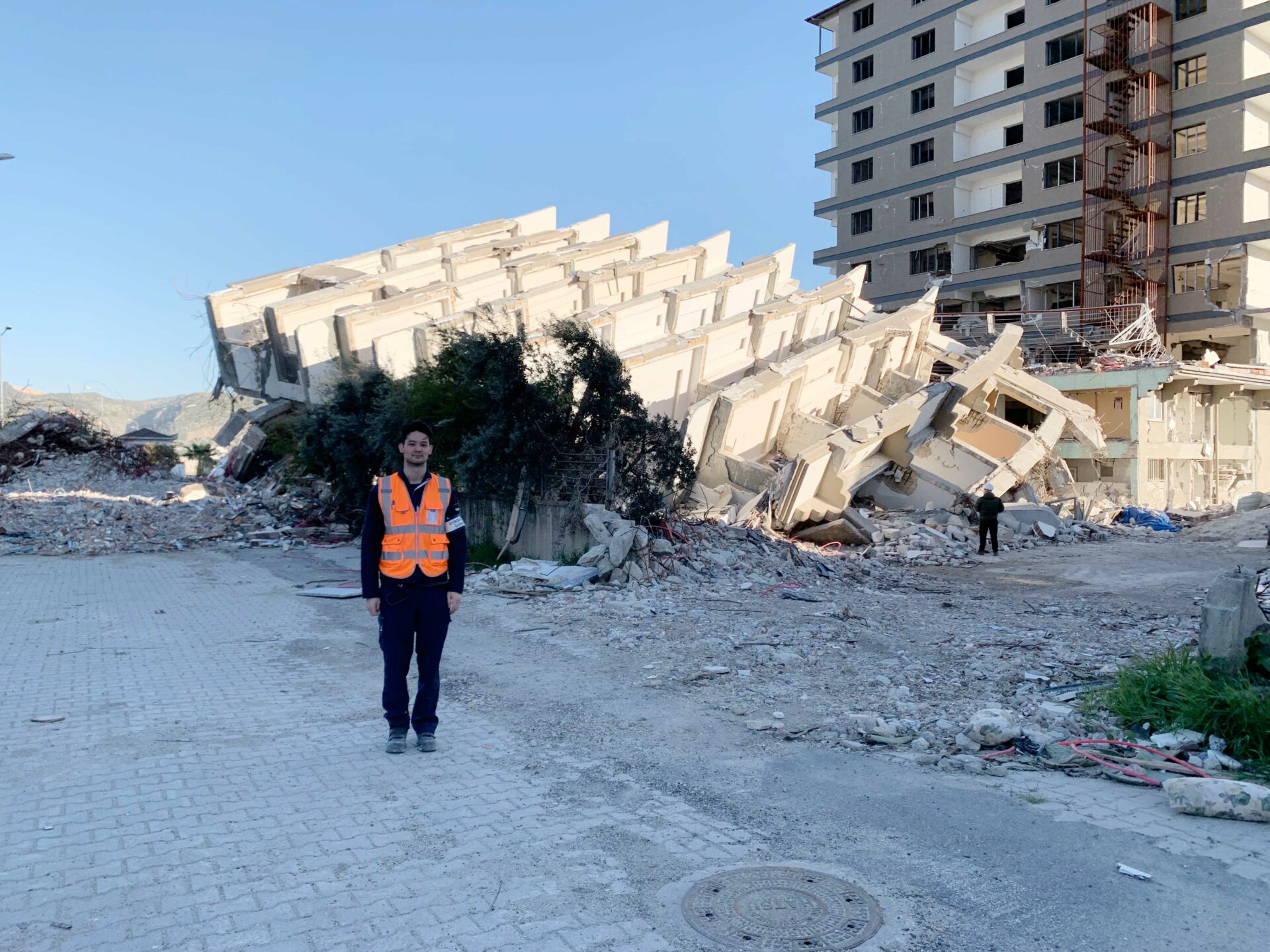 Alex standing in shadow, next to a multi-storey building that has toppled in the 2023 Turkey earthquake.